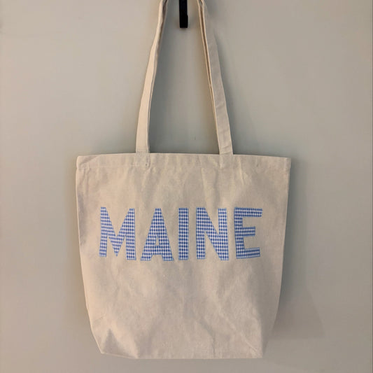 An off-white canvas tote bag with long handles, featuring the word 'MAINE' printed in blue gingham checkered letters on the front, hanging against a plain light gray wall.