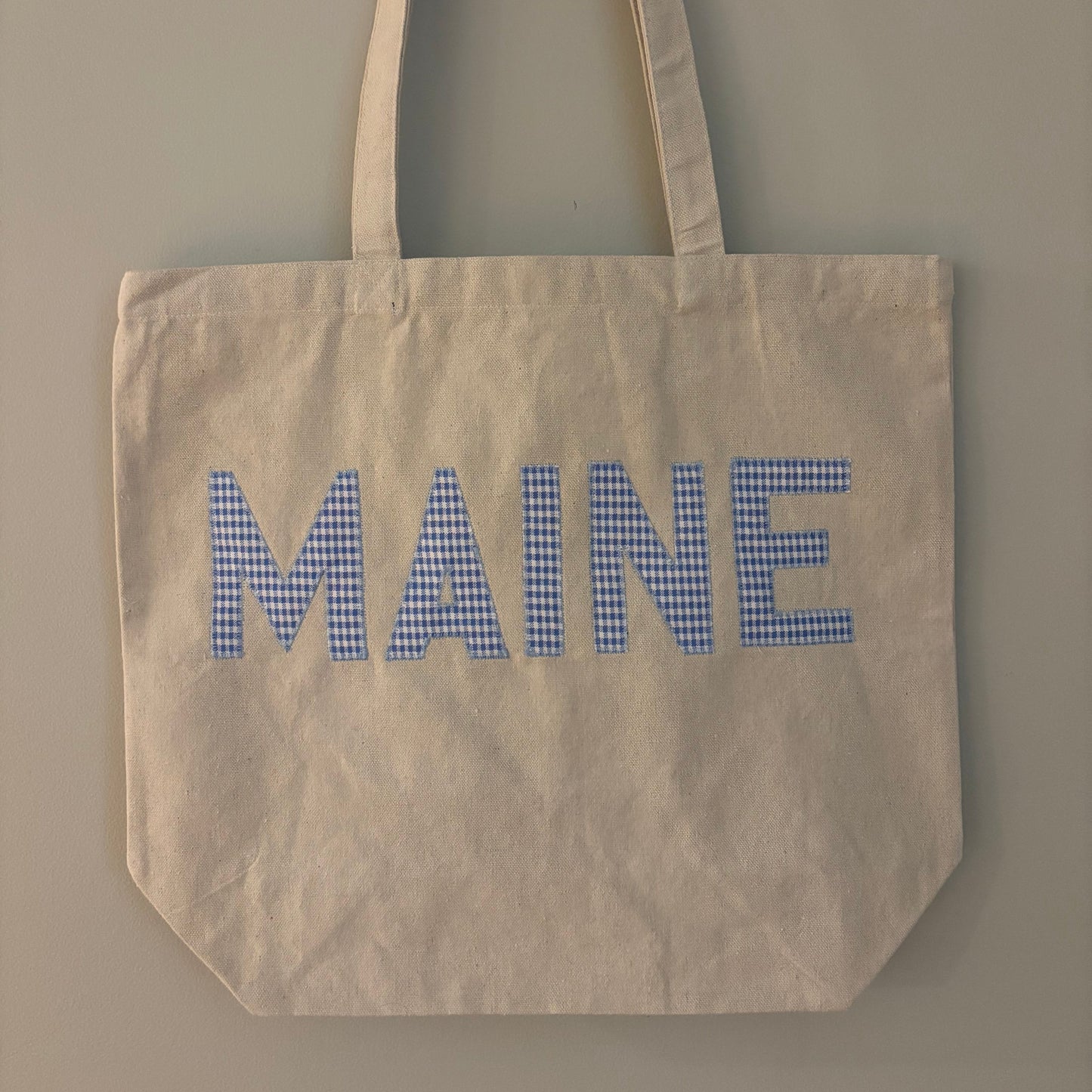 An off-white canvas tote bag with long handles, featuring the word 'MAINE' printed across the front in blue-and-white gingham checkered letters, hanging against a plain gray wall.