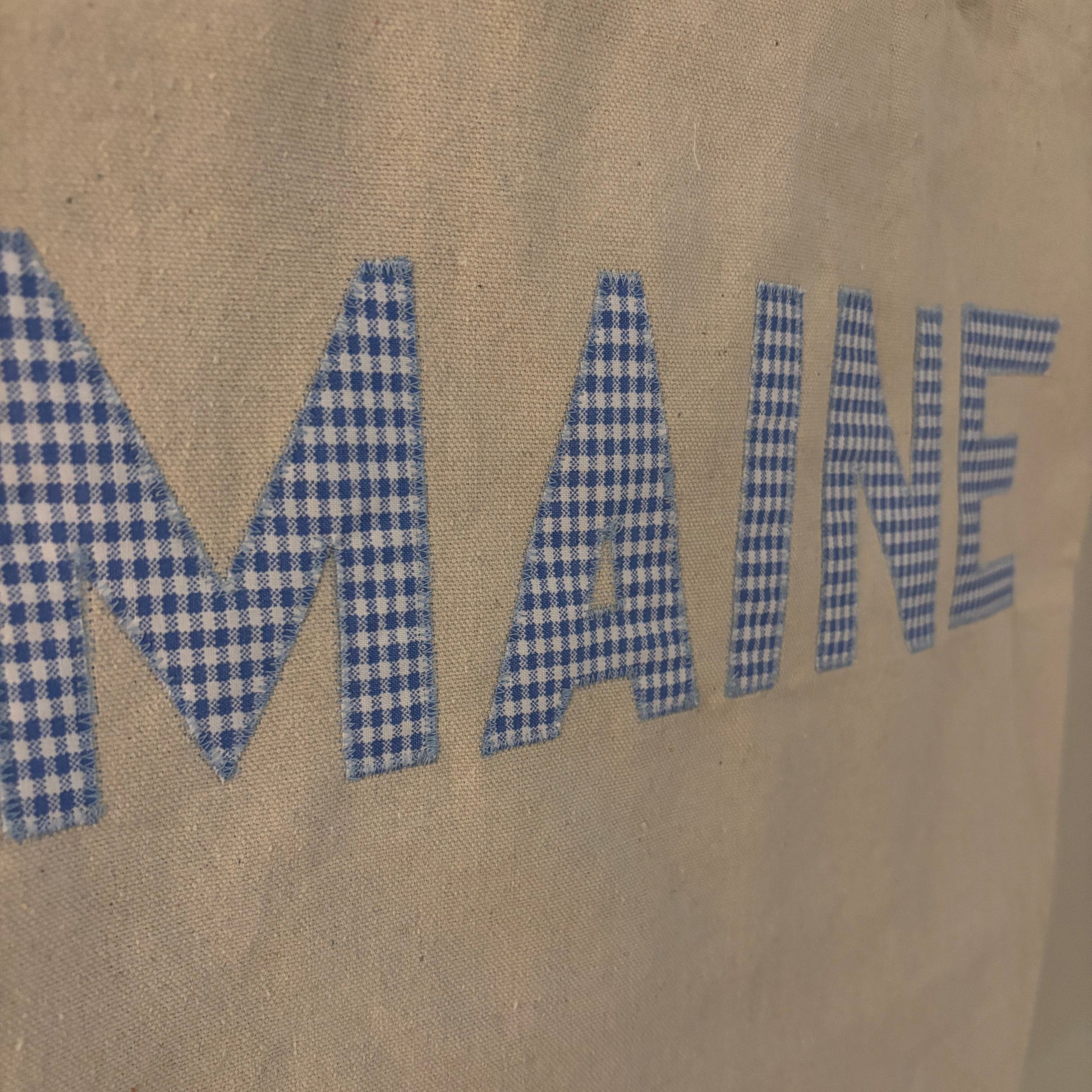 A close-up of a beige canvas tote bag featuring the word 'MAINE' in large blue-and-white gingham checkered letters.